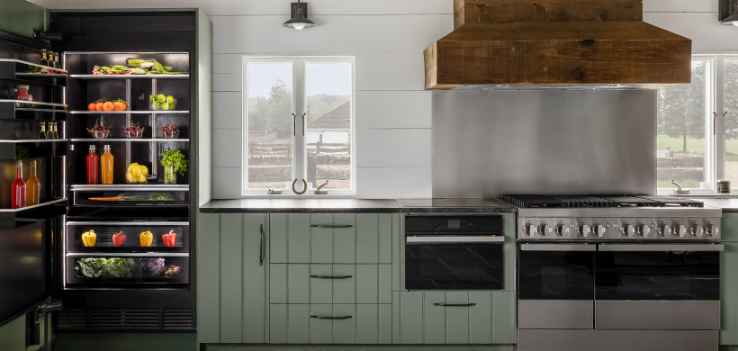 A kitchen with an opened JennAir Refrigerator filled with fresh herbs, beverages, produce and other food items. Also in the kitchen is a JennAir Double Oven below a JennAir Over-the-Range Hood and a JennAir Built-In Microwave.